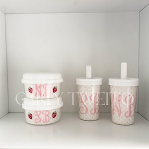 Op de afbeelding: Witte en roze drinkwaren en containers. Twee containers met deksel met aardbeienafbeeldingen en roze initialen. Twee bekers met witte deksels, rietjes, roze initialen en een stippenpatroon. De tekst "CARLY TWEITO" is zichtbaar.