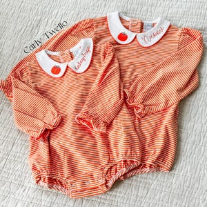 Puede incluir: Dos peleles de bebé a rayas naranjas y blancas con cuellos blancos. Cada cuello tiene una calabaza bordada en rojo y un nombre en escritura roja. Los peleles tienen mangas largas con puños con volantes.