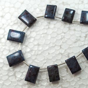 Puede incluir: Una hebra de cuentas de piedras preciosas de color azul oscuro y gris con un alambre plateado enhebrado. Las cuentas son de forma rectangular y tienen un patrón moteado.