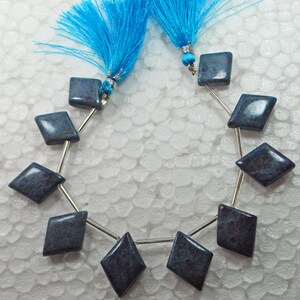 Puede incluir: Hilo de cuentas en forma de diamante en tonos azules y burdeos, ensartadas en alambre de plata. Las cuentas están pulidas y tienen un acabado brillante. Se ven dos borlas azules brillantes en la parte superior del hilo.