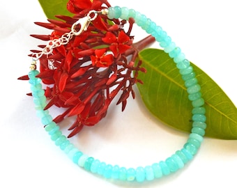 May include: A delicate bracelet featuring small, rounded, light blue beads. The bracelet is strung on a silver chain with a clasp. The jewelry is displayed with red flowers and a green leaf against a white background.