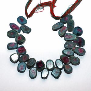 May include: A necklace of polished gemstones with a mix of teal and ruby red hues. The stones are strung together with red and green threads, creating a unique and colorful accessory. The gemstones have an oval and irregular shape.