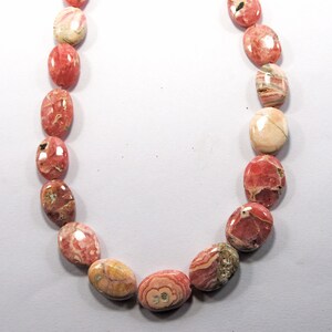 Puede incluir: Un collar de piedras preciosas ovaladas en tonos rosa, rojo y crema. Las piedras pulidas muestran patrones naturales. Las piedras están ensartadas para formar un bucle.
