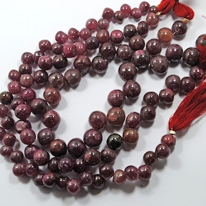 Op de afbeelding: Een streng diep rode edelsteen kralen met een kwast aan elk uiteinde. De kralen zijn rond en hebben een licht ruwe textuur.