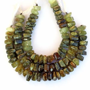 Pode incluir: Um fio de contas de pedras preciosas verdes e marrons. As contas são facetadas e têm forma retangular. As contas são enfiadas em um cordão branco.