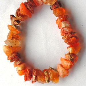 May include: A strand of carnelian gemstone beads in various shades of orange and red. The beads are irregular in shape and size, and are strung on a white cord.