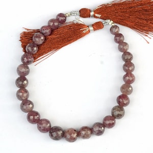 Può includere: Una collana di perline sfaccettate, rotonde e traslucide di colore viola rosato scuro disposte in forma circolare. La collana è fissata nella parte superiore con tappi in metallo argentato e presenta due lunghe nappe decorative in tessuto color ruggine.