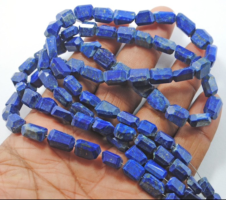May include: A strand of deep blue lapis lazuli beads, each bead is faceted and has a rectangular shape. The beads are strung on a wire and are ready to be used for jewelry making.