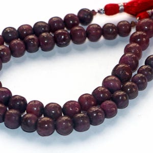 Op de afbeelding: Een streng donkerpaarse edelsteen kralen. De ronde kralen hebben een gepolijste afwerking. Rode kwasten met zilverkleurige accenten zijn aan de uiteinden bevestigd. De kralen zijn in een dubbele streng gerangschikt.