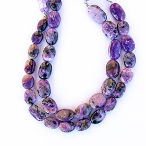 Puede incluir: Una hebra de cuentas de piedras preciosas de color púrpura y negro, con un borla en el extremo. Las cuentas son de forma ovalada y tienen un acabado natural y pulido.
