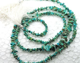 Può includere: Una collana di perline di turchese a forma di chip, con un fiocco bianco a un'estremità. Le perline sono un mix di turchese chiaro e scuro, e sono tutte di forma irregolare.