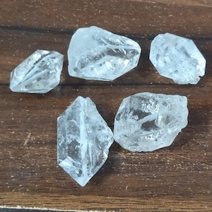 Puede incluir: Cinco cristales de cuarzo transparentes y en bruto con formas irregulares, multifacéticas y bordes afilados. Cada piedra muestra una apariencia translúcida similar al vidrio con fracturas internas naturales y formas geométricas variadas y rugosas.