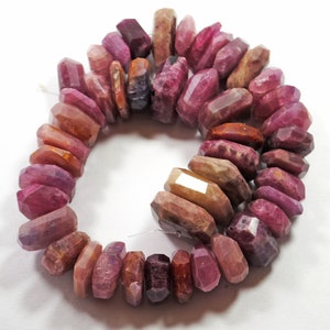 May include: A strand of pink, red, and brown faceted gemstone beads. The beads are all roughly the same size and shape, and they are strung together on a clear thread.