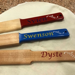 May include: Three wooden paddles with painted handles. One handle is red with the name "Svien" in blue, another is blue with "Swenson" in gold, and the third is natural wood with "Dyste" in red.