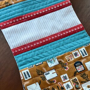 May include: A quilted table runner with a brown fabric featuring a repeating pattern of envelopes, pine branches, and the words "Air Mail" and "Mail". The runner is trimmed with teal blue fabric, red fabric with white stitching, and white and beige striped fabric.