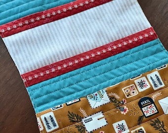 May include: A quilted table runner with a brown fabric featuring a repeating pattern of envelopes, pine branches, and the words "Air Mail" and "Mail". The runner is trimmed with teal blue fabric, red fabric with white stitching, and white and beige striped fabric.