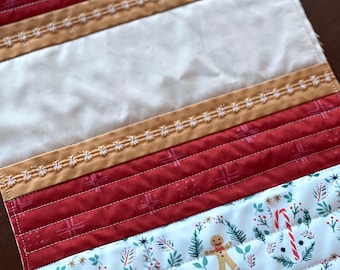 May include: A quilted table runner with a white background and a pattern of gingerbread men, candy canes, and evergreen trees. The runner has red and tan stripes with white stitching.