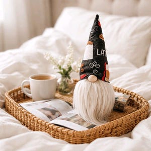 May include: A coffee-themed gnome with a black hat featuring coffee cup and bean designs, and the word "LATTE". The gnome has a white beard, a beige nose, and is on a woven tray with a cup of coffee and a magazine.