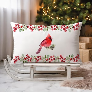 May include: A rectangular white throw pillow featuring a central watercolour illustration of a bright red cardinal bird perched on a pine branch, framed by horizontal borders of red holly berries and green leaves on the top and bottom edges.