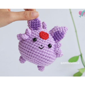 May include: A crocheted purple and red amigurumi toy resembling a Pokemon character. The toy has a heart-shaped nose and black eyes.