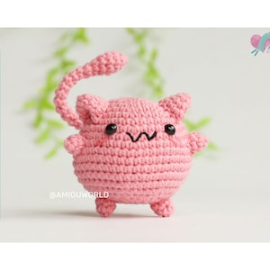 May include: A pink crocheted cat amigurumi toy with black eyes and a small smile. The toy has a curled tail and stands on four small legs.