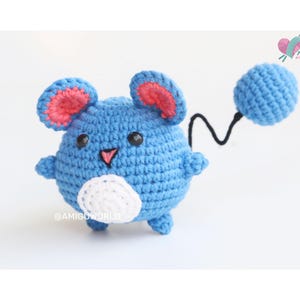 May include: A blue and white crocheted amigurumi toy shaped like a mouse with a long tail. The toy has pink ears and black eyes.