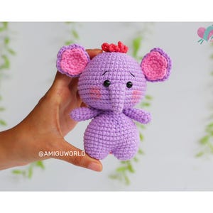 May include: A handmade purple crochet elephant with pink inner ears and a small red tuft of hair. The elephant has small black eyes and rosy cheeks. The image includes the text "PDF PATTERN" and the Instagram handle "@AMIGUWORLD".