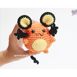 Könnte beinhalten: Eine gehäkelte orangefarbene und schwarze Pokémon-Figur mit gelben Akzenten. Die Figur hat große Ohren und einen kleinen Schwanz. Die Figur wird von einer Hand gehalten.