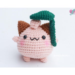 May include: A crocheted amigurumi toy in the shape of a pink creature with brown ears and a green leaf on top. The toy has a cute face with black eyes and a small mouth.
