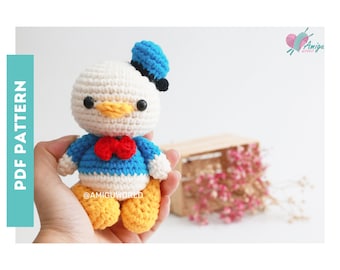 Op de afbeelding: Gehaakt amigurumi speelgoed van Donald Duck, een cartoonfiguur. Het speelgoed is wit met een gele snavel, een blauw shirt, een rode strik en gele benen. Het speelgoed draagt een blauw-zwarte matrozenhoed.