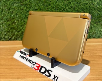 Peut inclure: Console de jeu portable Nintendo 3DS XL dorée, posée sur un support blanc. Le support affiche "new Nintendo 3DS XL". Le dessus de la console présente un motif géométrique. Fond vert.