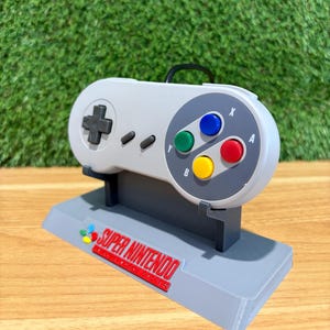 May include: A grey Super Nintendo controller replica on a black stand. The controller has coloured buttons and a directional pad. The base of the stand displays the words "Super Nintendo" in red.