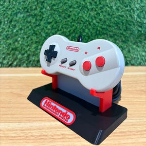 May include: A gray Nintendo controller with red and black accents, resting on a black stand with a red support. The stand features the Nintendo logo in red and white. The controller has a directional pad, buttons, and the text "SELECT START".