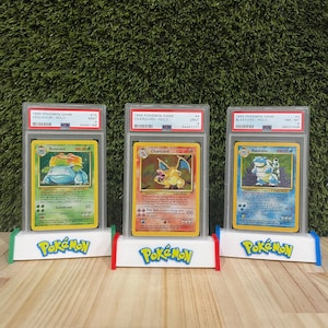 May include: Three graded Pokémon cards on custom stands. The cards feature Venusaur, Charizard, and Blastoise, with the text "1999 Pokémon Game" visible. Each card is encased in a protective holder with grading information.