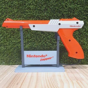 Op de afbeelding: Een oranje en witte Nintendo Zapper lichtpistool, een retro videogame-accessoire. Het pistool staat op een grijze standaard met de woorden "Nintendo Zapper" erop gedrukt. Het woord "Nintendo" staat ook op het pistool.