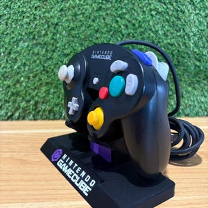 May include: Black Nintendo GameCube controller with colourful buttons and a white analogue stick. The controller rests on a black stand with the words "NINTENDO GAMECUBE" printed in white. The controller has a black cord.