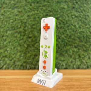 Può includere: Un telecomando Wii bianco con un pannello laterale verde e pulsanti arancioni, in una base di ricarica bianca. Il telecomando presenta un pad direzionale, un pulsante A e pulsanti numerati. Il logo Wii è visibile.