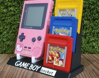 May include: A pink Game Boy Pocket console with a Hello Kitty design, displayed with three Pokémon game cartridges in a black stand. The cartridges are red, blue, and yellow.