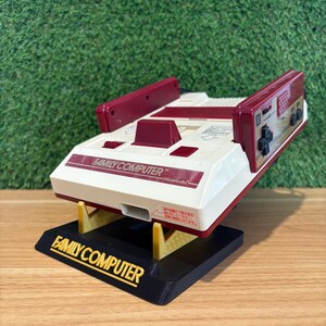 May include: A retro Family Computer games console in cream, burgundy, and black. The console is mounted on a black base with "FAMILY COMPUTER" in gold lettering. The Nintendo logo is visible. The console has two burgundy controller ports.