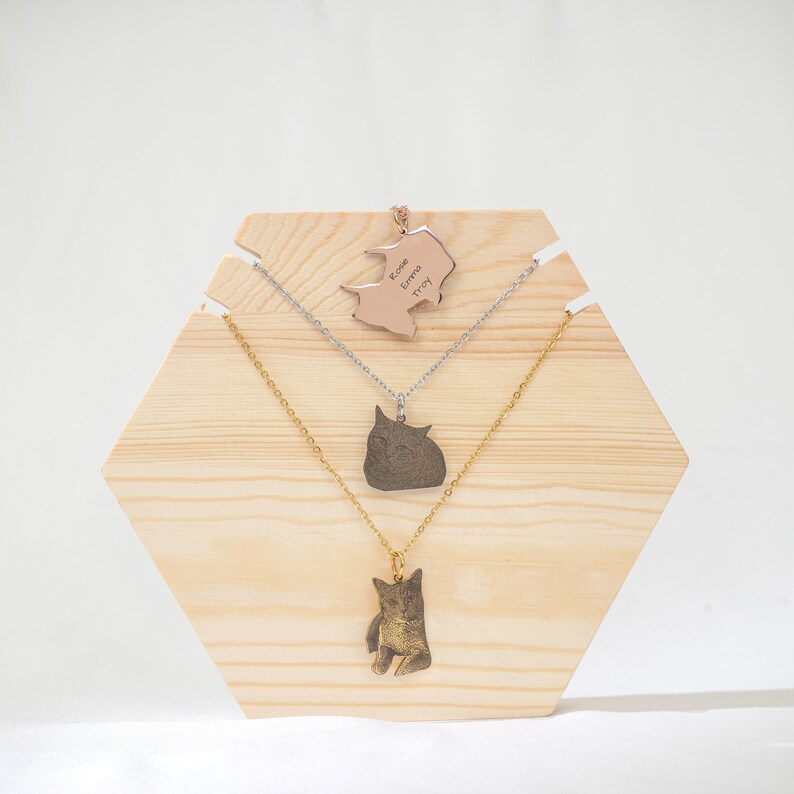 May include: Three necklaces displayed on a wooden hexagonal stand. The top necklace features a rose gold pendant with the names "Rose Emma Troy" engraved on it. The middle necklace has a silver pendant with a cat silhouette. The bottom necklace has a gold pendant with a cat silhouette.