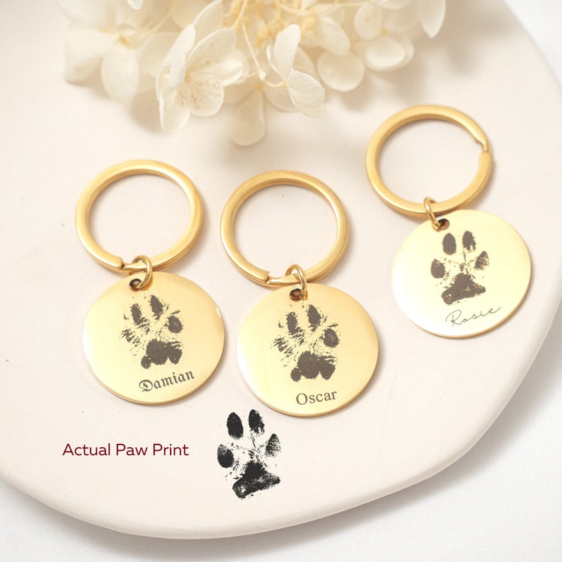 May include: Three gold keychains with engraved paw prints and names: Damian, Oscar, and Rosie. An actual paw print is shown below the keychains.