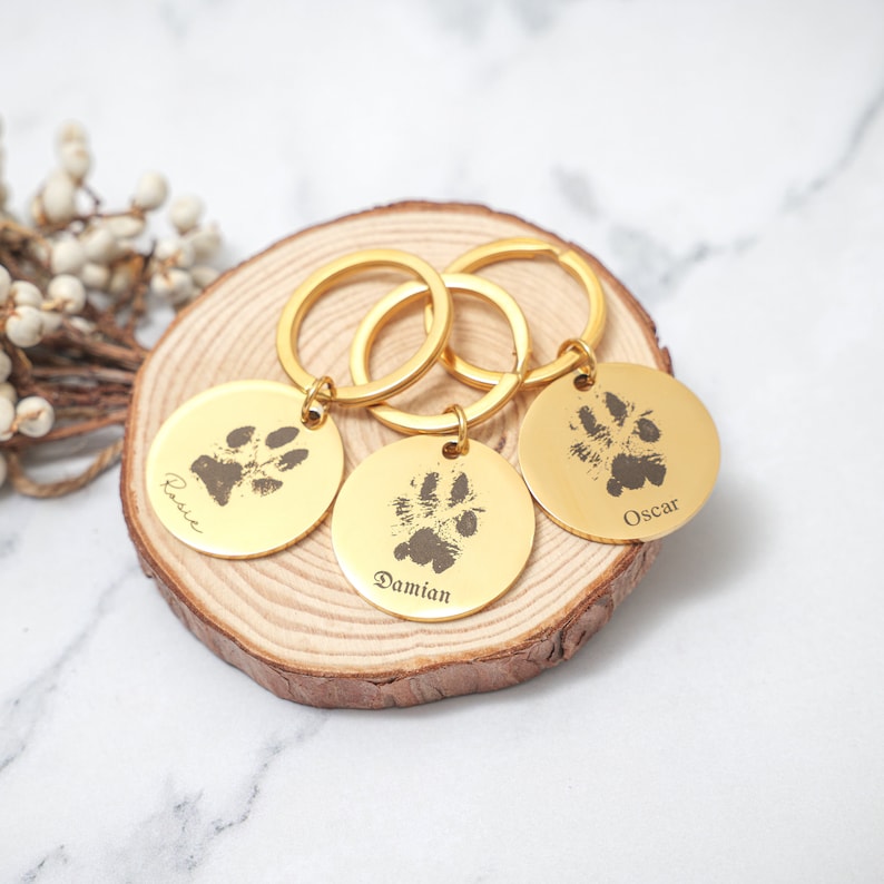 May include: Three gold keychains with engraved paw prints and names: Rosie, Damian, and Oscar. The keychains are on a wooden surface.