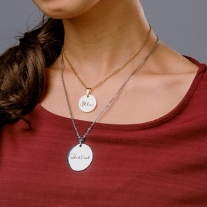 Può includere: Due collane d'argento con ciondoli rotondi. Il ciondolo superiore è bianco con la parola "Elle" scritta in caratteri dorati. Il ciondolo inferiore è bianco con il testo "Sophia & Kenneth" scritto in caratteri neri.