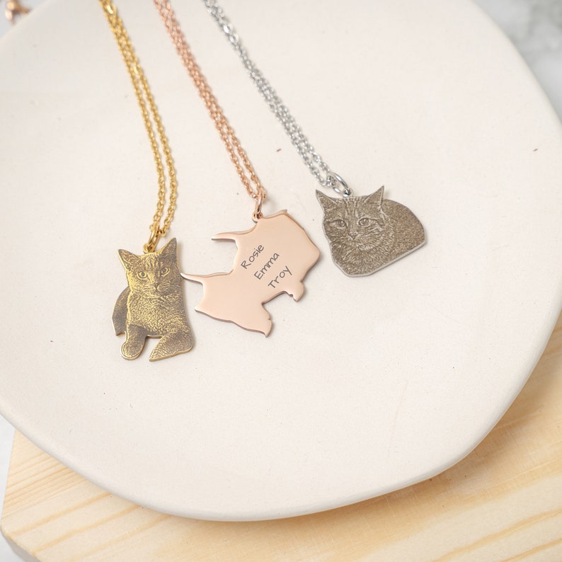May include: Three silver necklaces with personalized cat photo pendants. The pendants are in the shape of a cat, a heart, and a paw print. The necklaces are on a white surface.