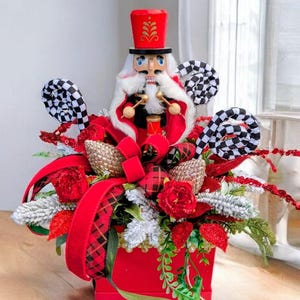 May include: A Christmas-themed centrepiece with a wooden nutcracker soldier in a red hat and coat, surrounded by red roses, tartan ribbons, and decorative elements. The arrangement is in a red square box.