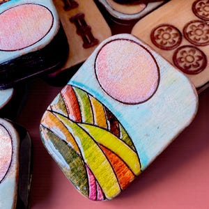 Handmade wooden Riichi Mahjong tiles featuring a colorful sunrise design with pink sun and rolling hills.