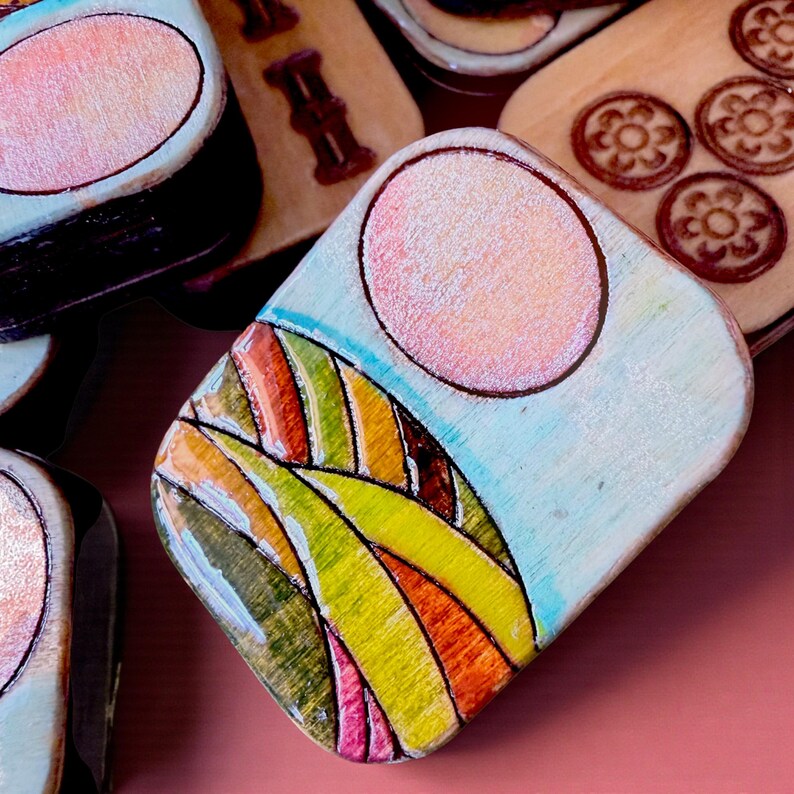 Handmade wooden Riichi Mahjong tiles featuring a colorful sunrise design with pink sun and rolling hills.
