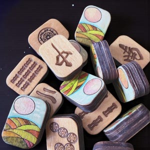 Handmade wooden Riichi Mahjong tiles featuring a colorful sunrise design with pink sun and rolling hills.