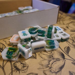 Pu&ograve; includere: Una collezione di tessere Mahjong fatte a mano. Le tessere sono bianche con dettagli verdi e dorati, alcune con caratteri cinesi. Sono disposte su una superficie a motivi floreali, con una scatola sullo sfondo.