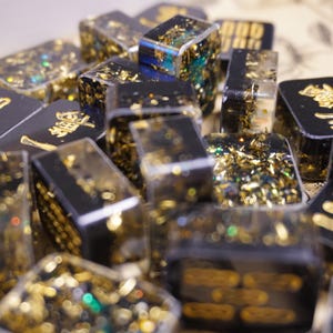 Op de afbeelding: Een verzameling zwarte en heldere harsblokken met gouden vlokken en gouden letters. De blokken zijn rechthoekig en hebben een glanzende afwerking. Sommige blokken hebben gouden Chinese karakters.